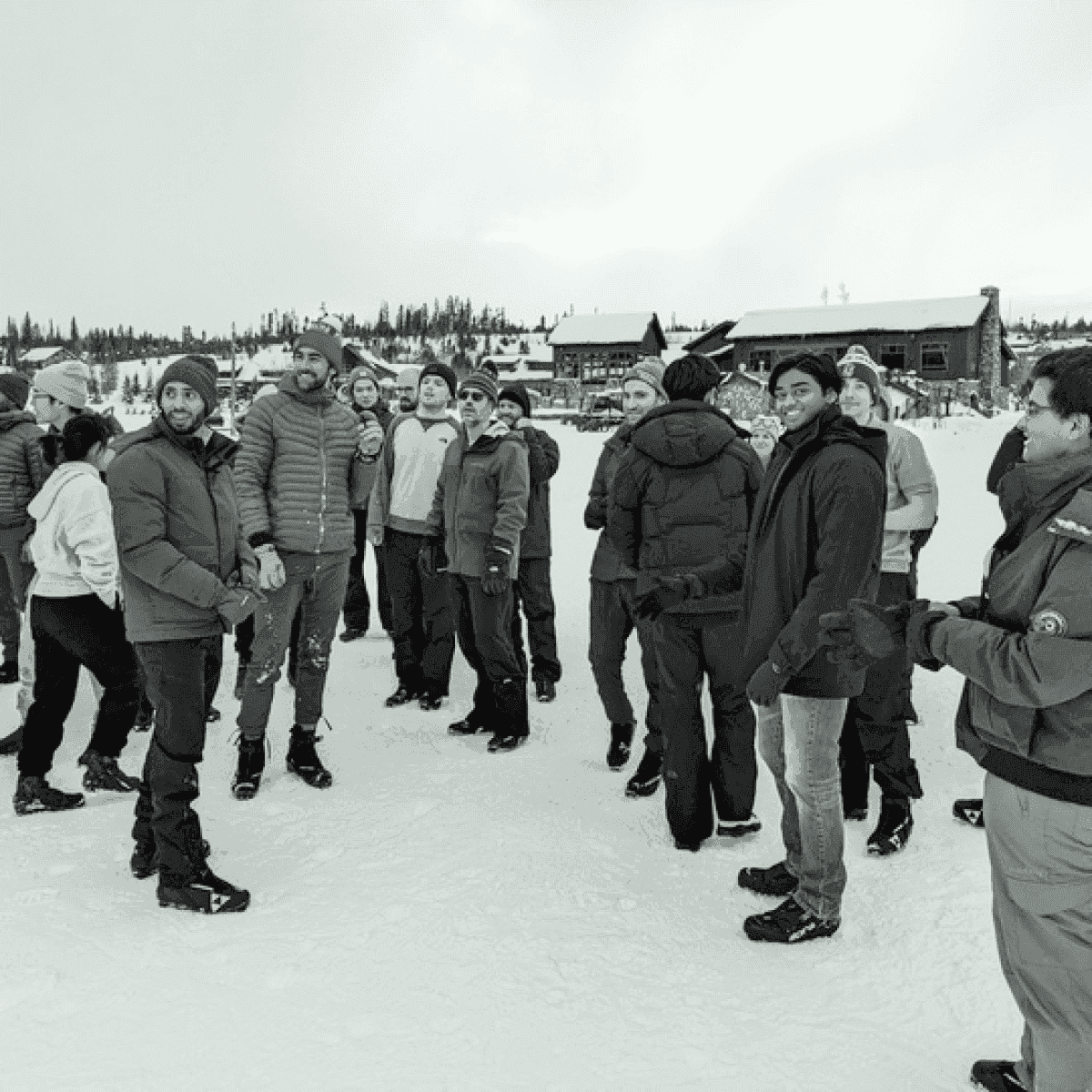 A diverse group of people in winter clothing stand in the snow, with buildings and trees in the background.